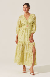 A woman is wearing a light yellow, embroidered maxi dress with a V-neckline. The dress features long, puffy sleeves, a gathered waist, and a high slit on one side revealing her leg. She is pairing the outfit with white open-toe heels.